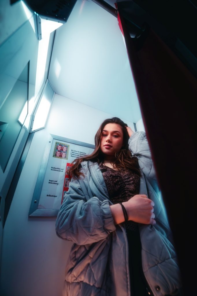 Zu sehen ist ein Porträt aufgenommen in München in Bayern. Im Porträt Foto ist eine junge Frau zu sehen, zum Teil auch Neon-Lichter, eine U-bahn, Fotoautomat und Rolltreppe. Die Aufnahmen an der Oberfläche wurden in München, nähe Rotkreuzplatz aufgenommen. Im Hintergrund der Porträts sind eine Schule und ein Krankenhaus zu sehen. Die Bilder sind für eine Portfolio Erweiterung geschossen worden.