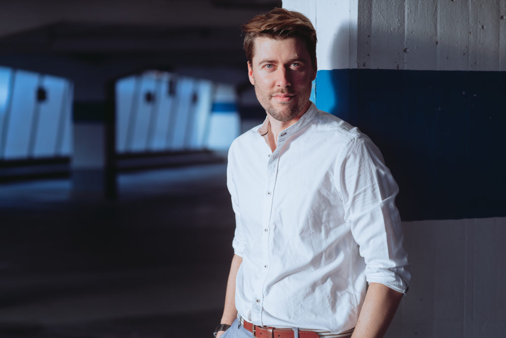 Das etwa 1,80 Meter große, brünette männliche Model Axel posiert in einem Parkhaus in der Abendsonne bei einem Shooting mit dem Münchner Fotograf Daniel Schubert. Das Bild wird auch als Beispiel für Fotografie Kurse oder Portrait Workshops benutzt.